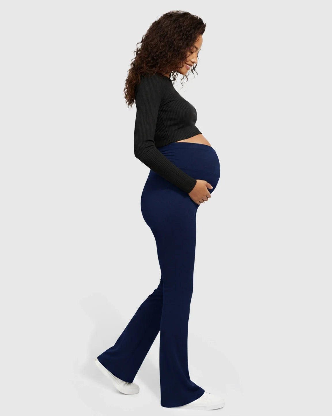 Pregnant woman in navy Bump Society flared maternity pants with a crossed waistband, holding her bump.