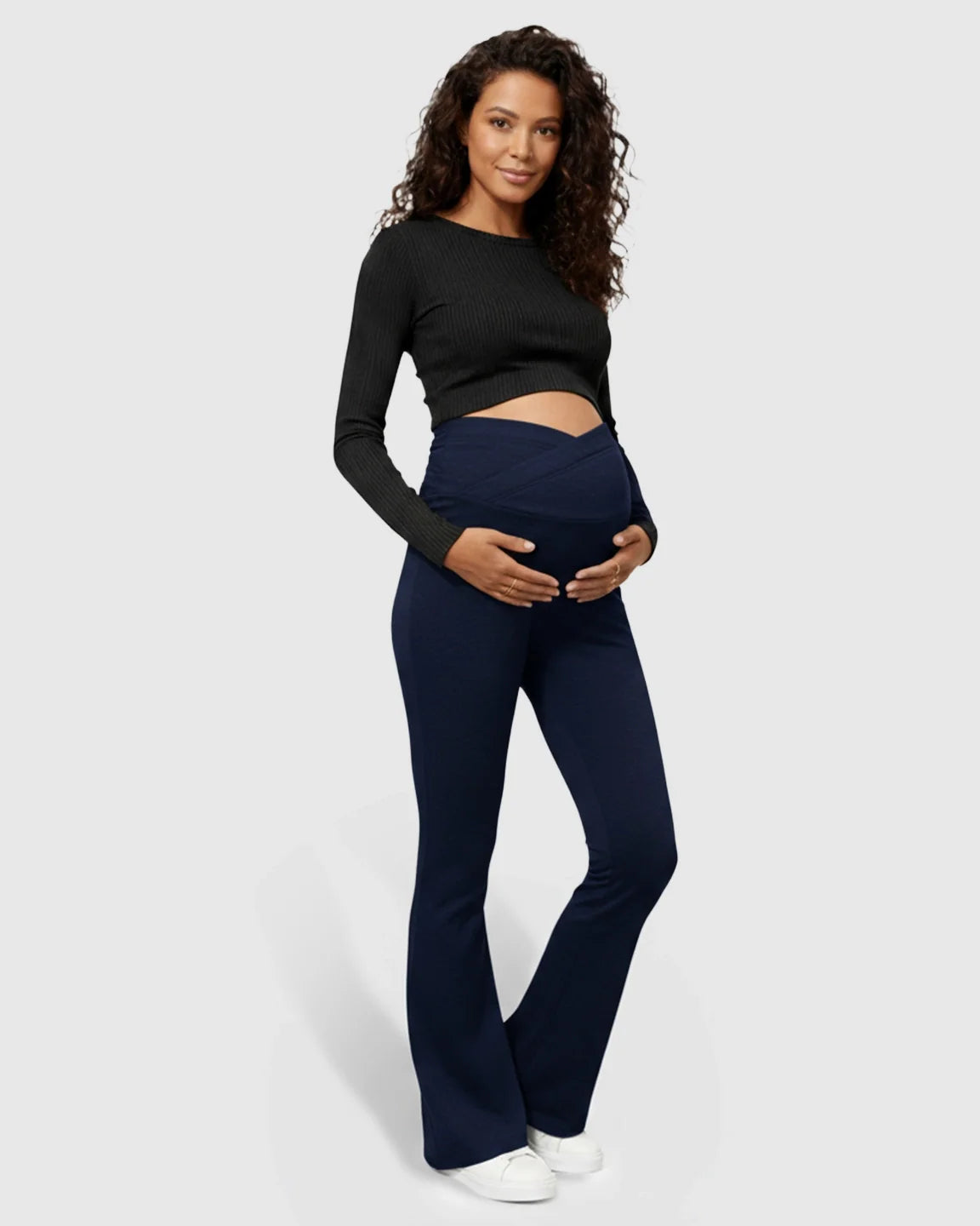 Maternity model in navy flared pants with a crossed waistband and black long-sleeve top, cradling her bump.