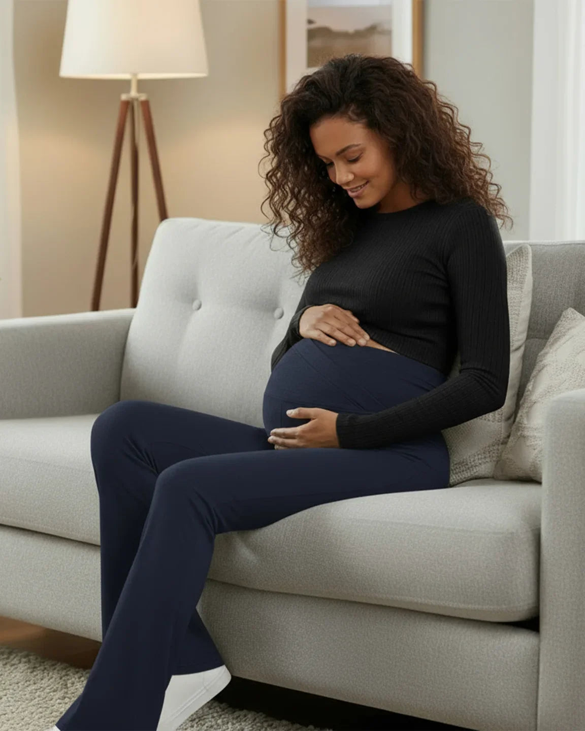Smiling pregnant woman in navy crossed waistband flared maternity pants and black top, sitting on a gray couch.
