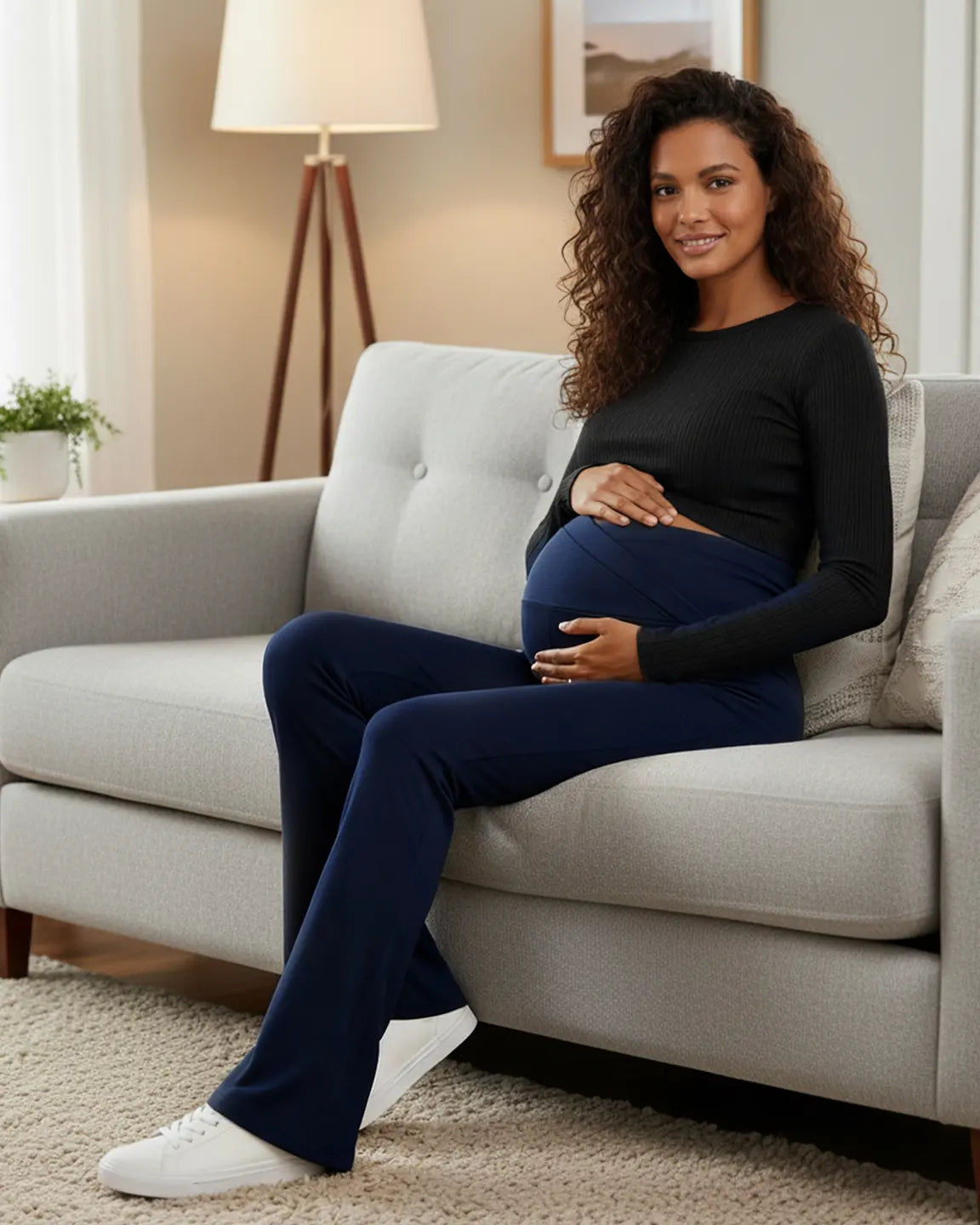 Smiling pregnant woman in navy crossed waistband flared maternity pants and black top on a gray couch.