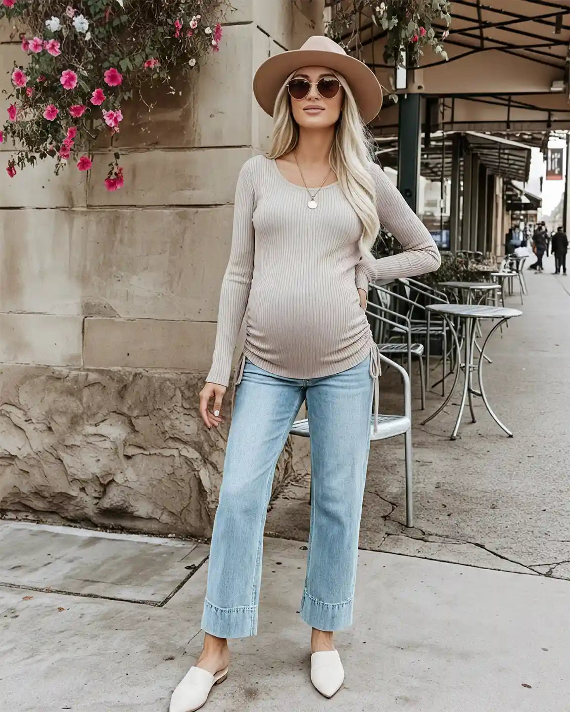 Blonde pregnant woman in a beige ribbed maternity top with adjustable side drawstrings, light wash jeans, and a hat.