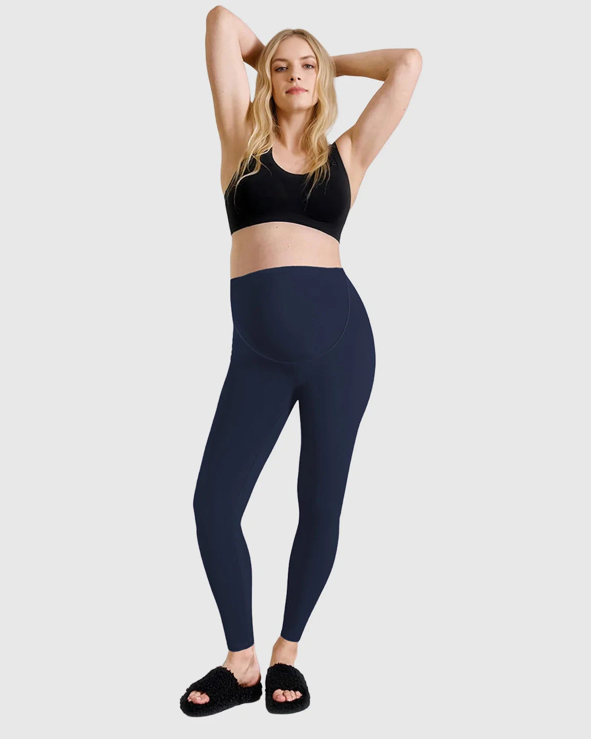 Woman wearing navy high-waist sculpting maternity leggings and a black sports bra, hands behind head.