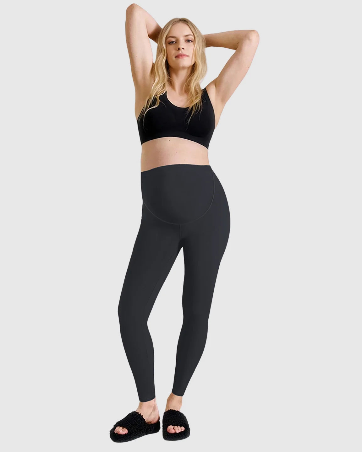 Blonde woman wearing black high-waist sculpting maternity leggings and a black sports bra, hands behind head.