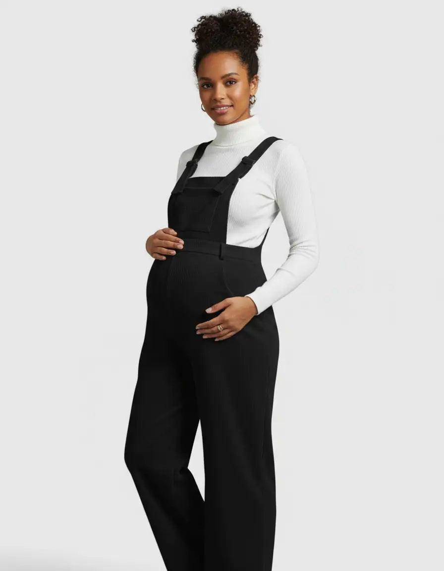 Smiling pregnant woman in a black corduroy maternity jumpsuit over a white turtleneck.