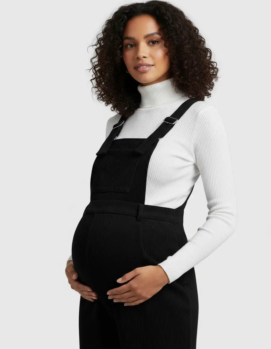 Pregnant woman in a black corduroy maternity jumpsuit with adjustable straps over a white turtleneck.