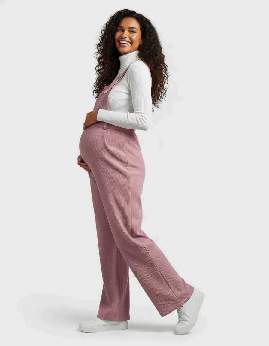 Smiling pregnant woman in a pink corduroy maternity jumpsuit with adjustable straps and a white turtleneck.