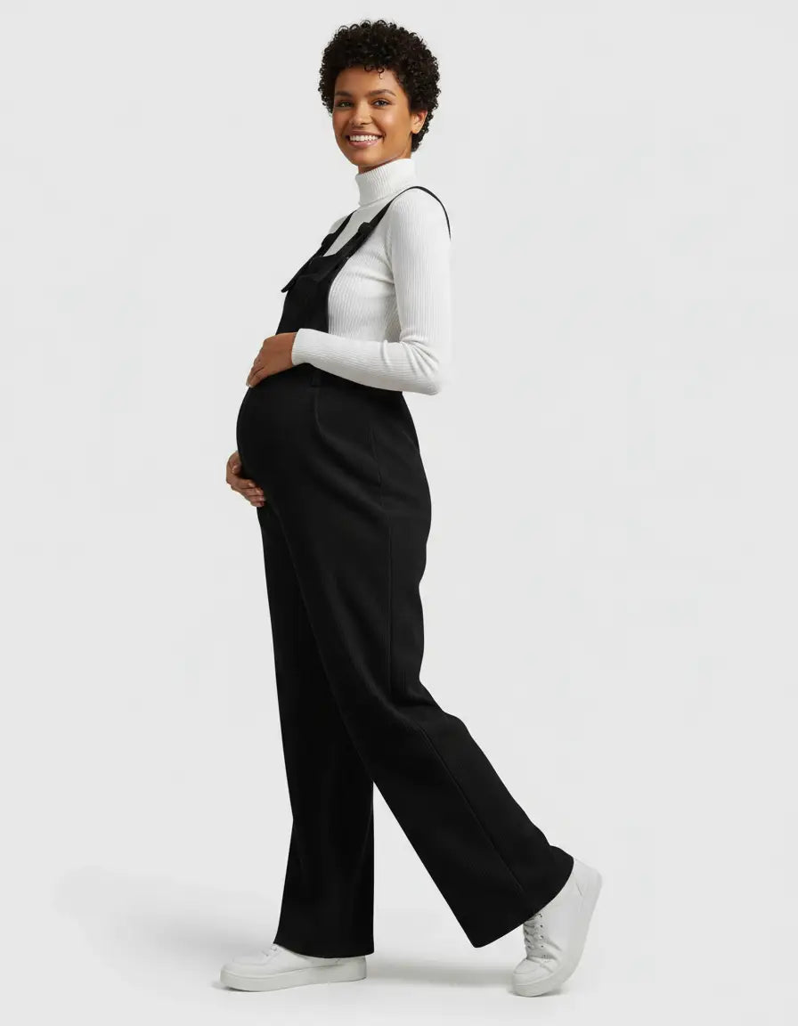 Smiling pregnant woman in a black corduroy maternity jumpsuit over a white turtleneck, holding her belly.