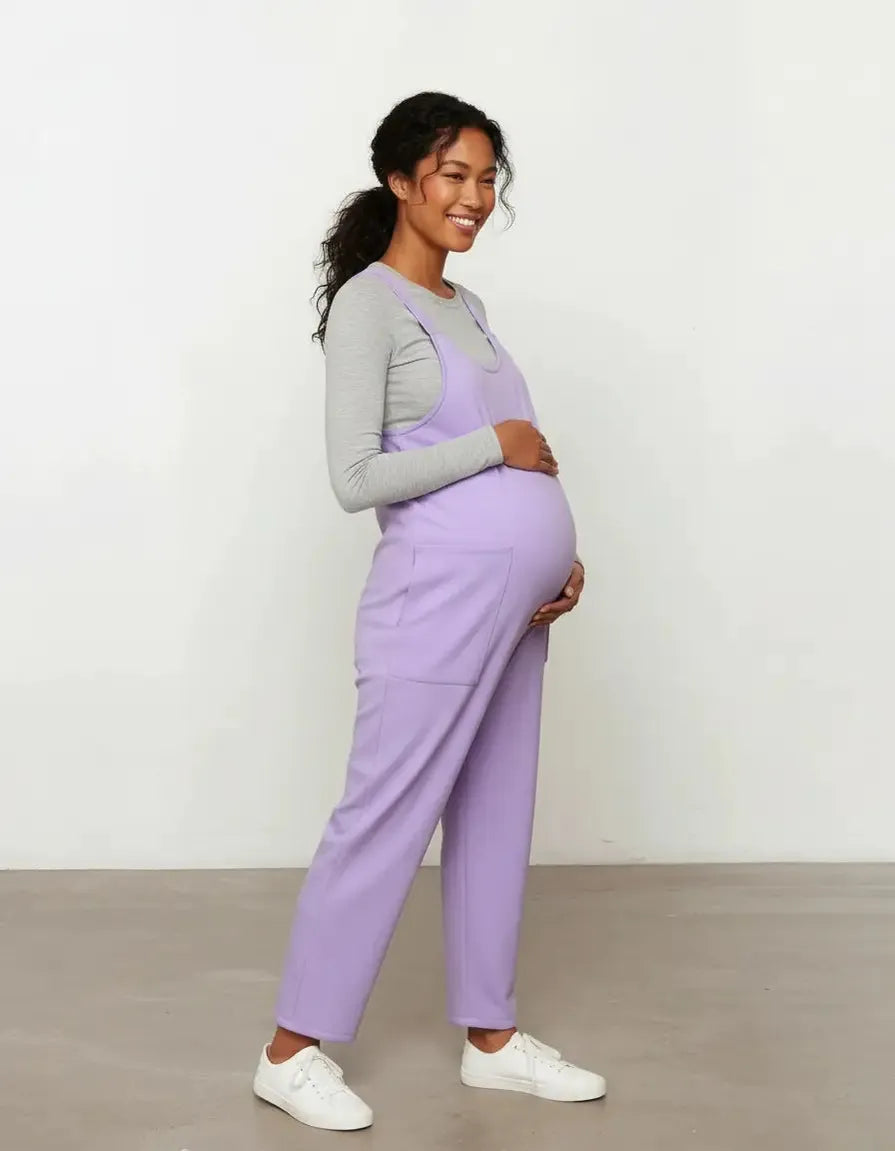 Smiling pregnant woman in a lavender flowy maternity jumpsuit with adjustable straps and pockets.