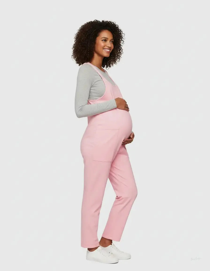 Smiling pregnant woman in a pink flowy maternity jumpsuit with adjustable straps and a gray long-sleeve top.