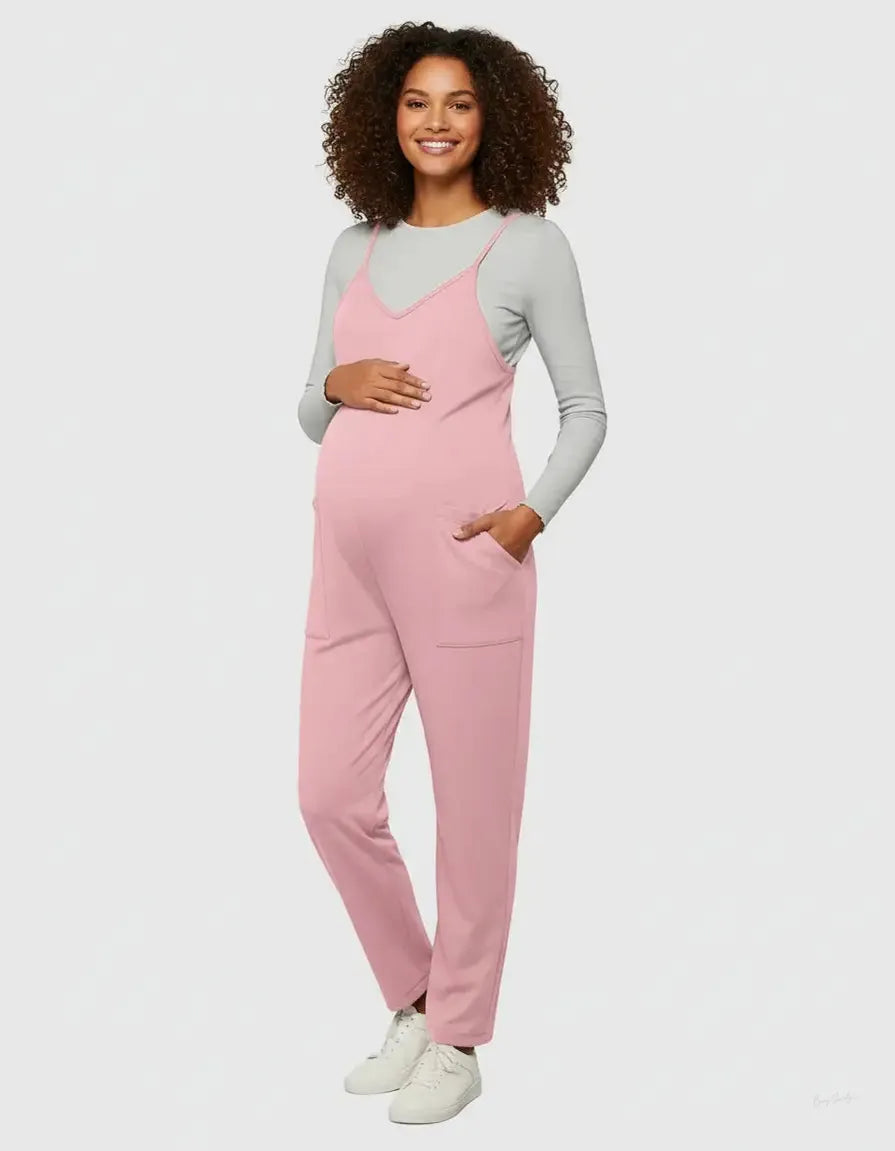 Pregnant woman in a pink flowy maternity jumpsuit with adjustable straps and pockets, over a grey long-sleeve shirt.