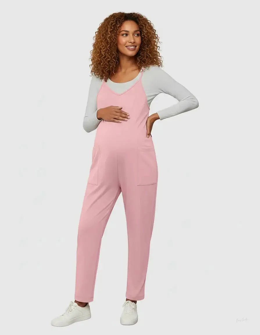 Smiling pregnant woman in a pink flowy maternity jumpsuit with adjustable straps and pockets, over a grey long-sleeve top.