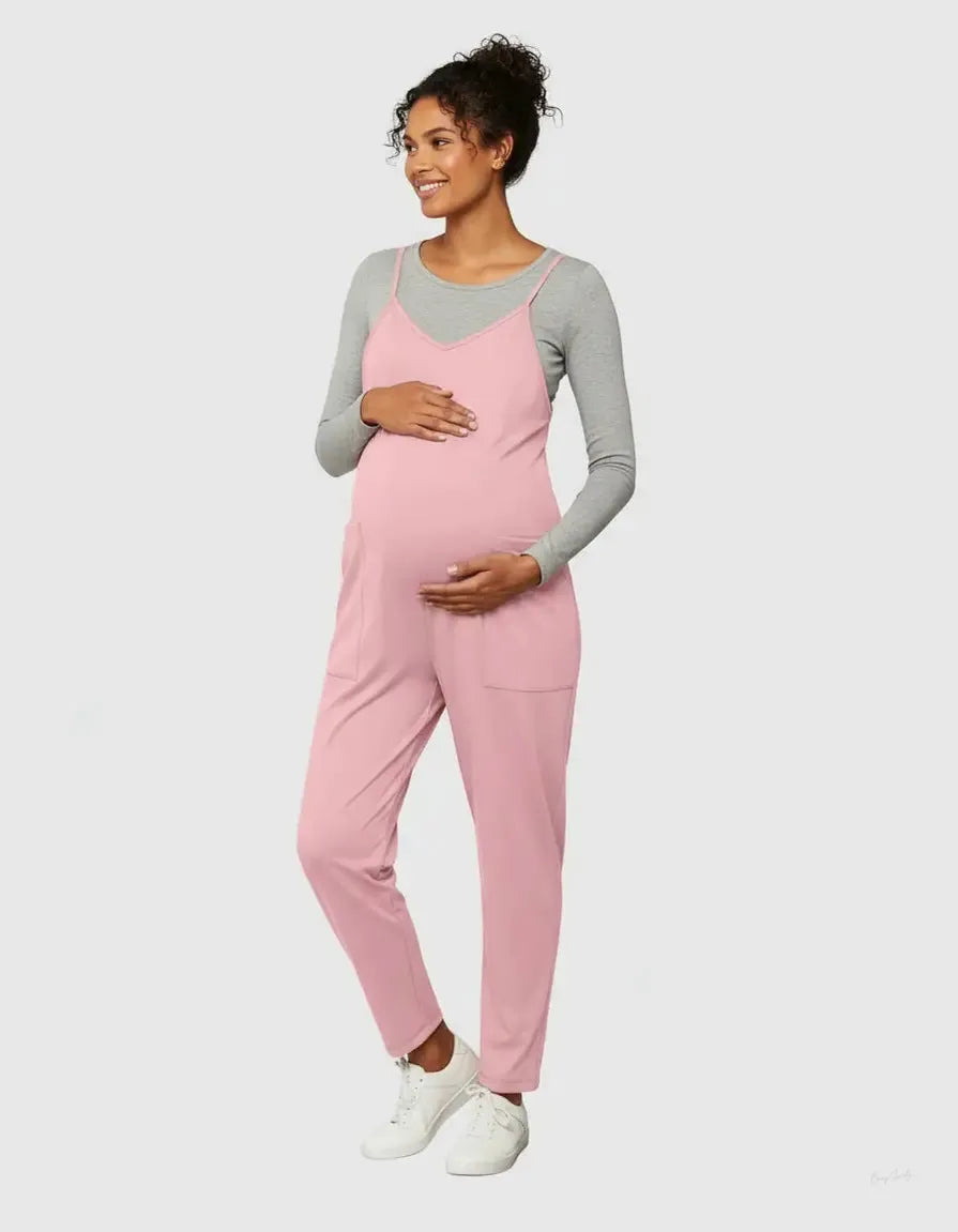Smiling pregnant woman in a pink flowy maternity jumpsuit with adjustable straps over a grey long-sleeve top.