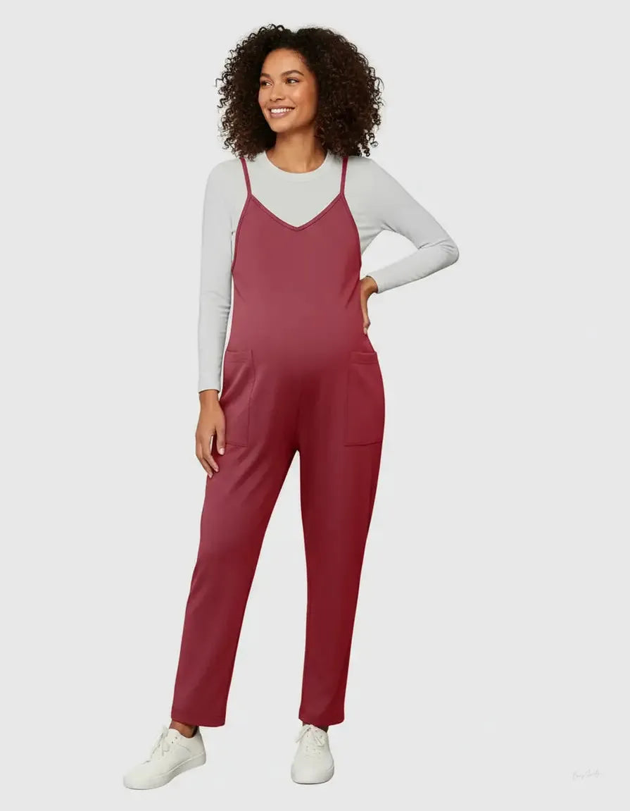 Smiling pregnant woman in a flowy, red maternity jumpsuit with adjustable straps over a white long-sleeve tee.