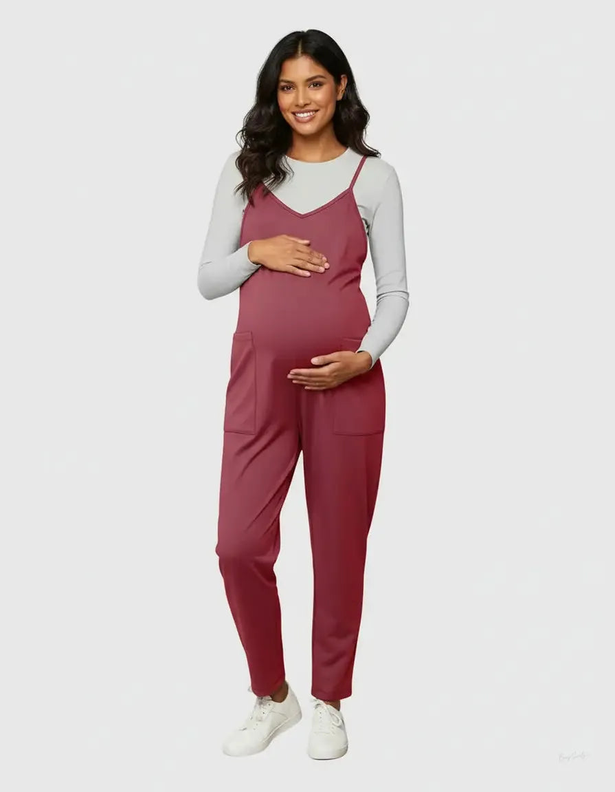 Smiling pregnant woman in a flowy, rose-colored maternity jumpsuit with adjustable straps and pockets, over a light grey long-sleeve top.