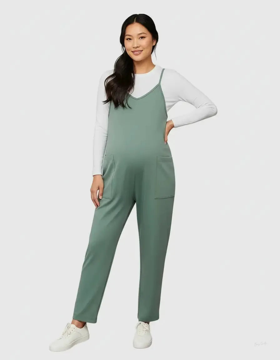 Woman in a sage green flowy maternity jumpsuit with adjustable straps, pockets, and a white long-sleeve shirt.