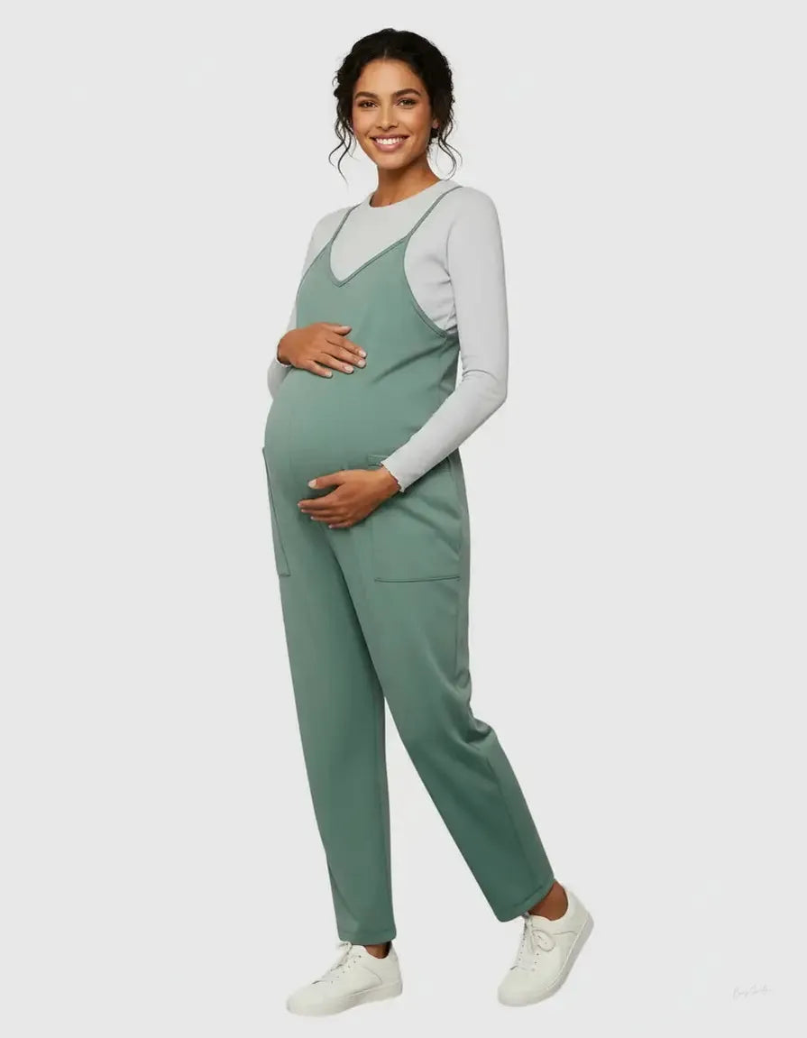Smiling pregnant woman in a sage green flowy maternity jumpsuit with adjustable straps and pockets, over a white long-sleeve top.
