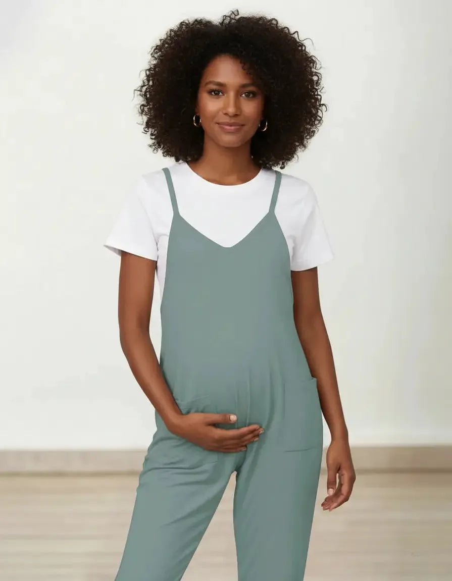 Maternity model in a sage green flowy jumpsuit with adjustable straps over a white tee, hands on bump.
