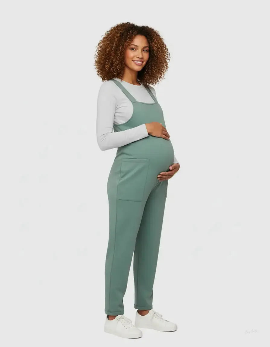 Smiling pregnant woman in a sage green flowy maternity jumpsuit with adjustable straps and pockets.