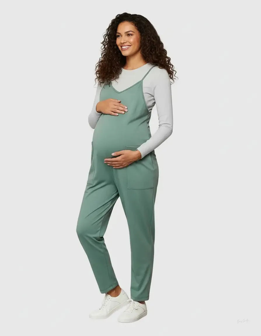 Pregnant woman in a sage green flowy maternity jumpsuit with adjustable straps and pockets, over a grey long-sleeve top.