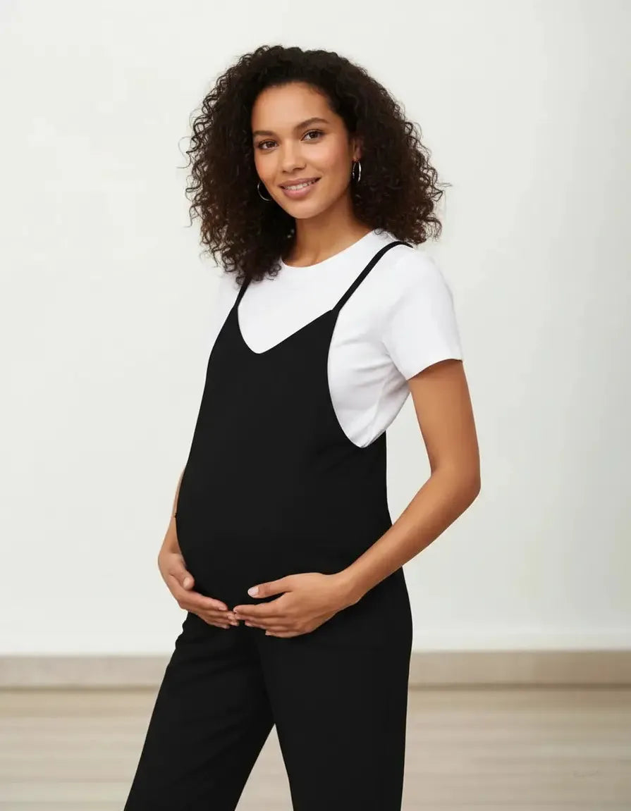 Smiling pregnant woman in a black flowy maternity jumpsuit with adjustable straps over a white tee.