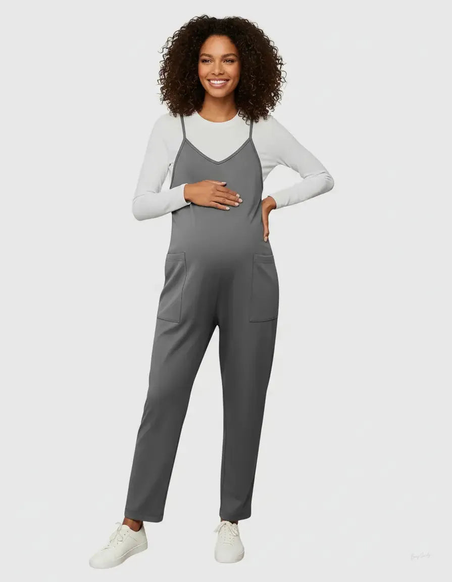 Smiling pregnant woman in a gray flowy maternity jumpsuit with adjustable straps and pockets, over a white long-sleeve shirt.