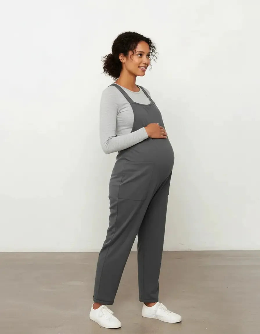 Pregnant woman in a charcoal gray flowy maternity jumpsuit with adjustable straps, over a light gray long-sleeve top.
