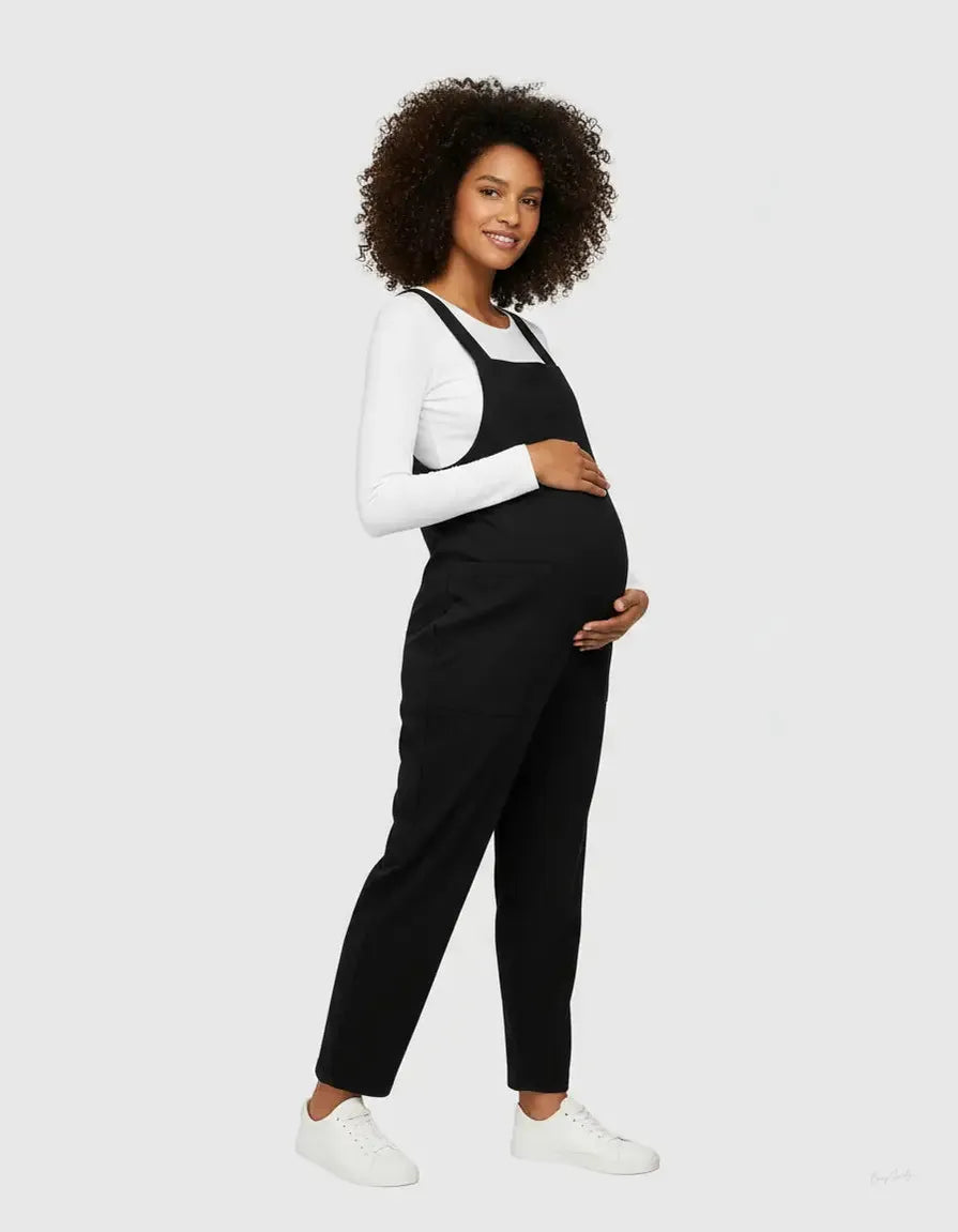 Smiling pregnant woman in a black flowy maternity jumpsuit with adjustable straps over a white long-sleeve top.