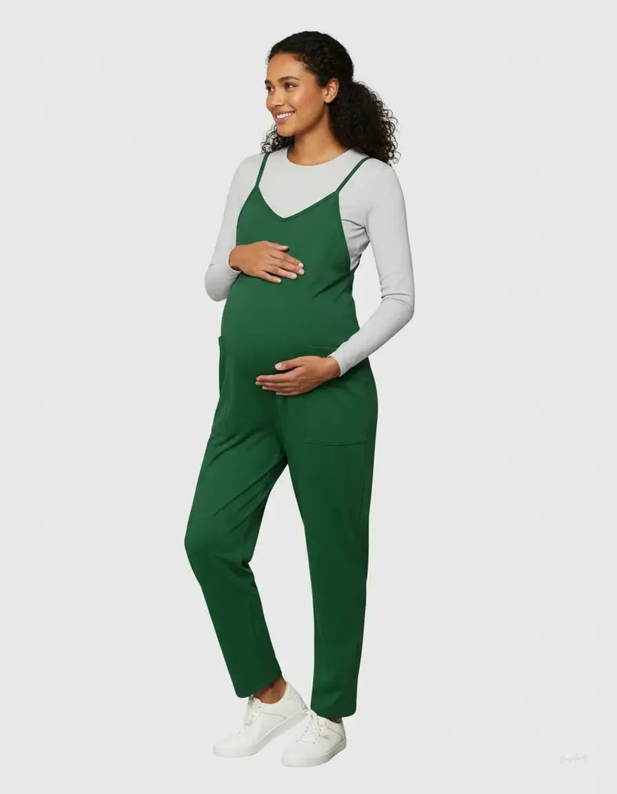 Smiling pregnant woman in a dark green adjustable strap flowy maternity jumpsuit with pockets over a light grey long-sleeve top.