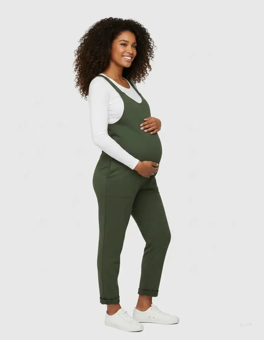 Smiling pregnant woman in a dark green flowy maternity jumpsuit with adjustable straps over a white long-sleeve top.