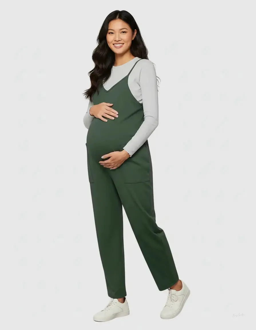 Smiling pregnant woman in a dark green maternity jumpsuit with adjustable straps and pockets, over a light grey long-sleeve top.