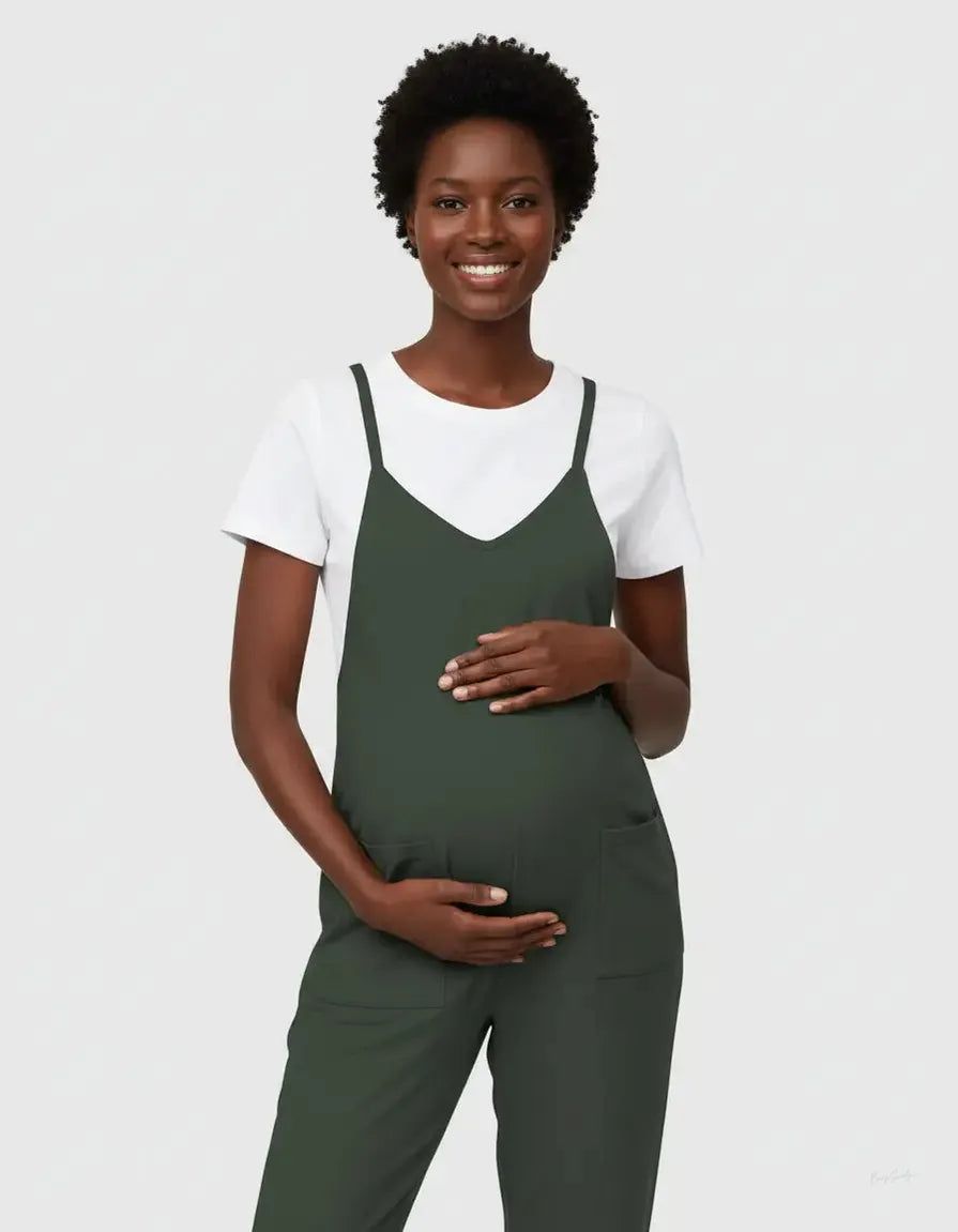Smiling pregnant woman in a dark green flowy maternity jumpsuit with adjustable straps over a white tee.