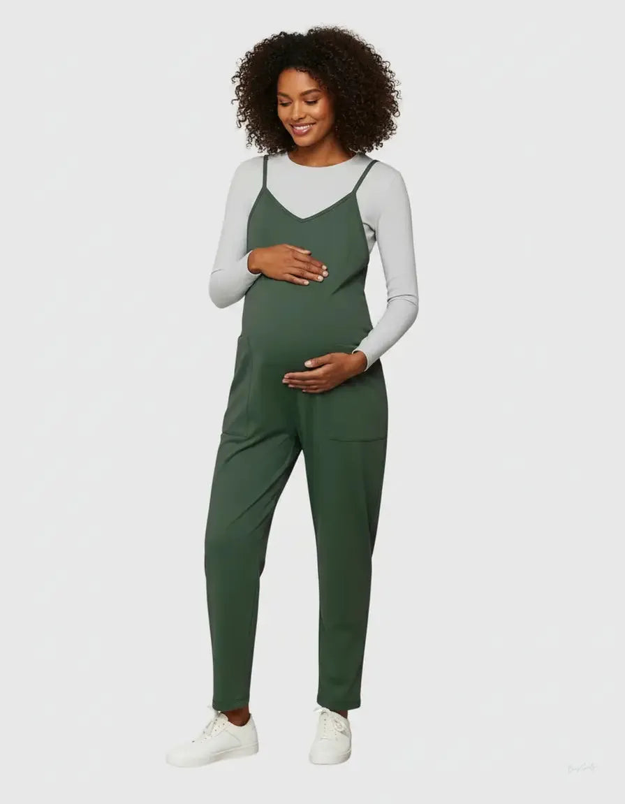 Smiling pregnant woman in a dark green adjustable strap flowy maternity jumpsuit over a white long-sleeve top.