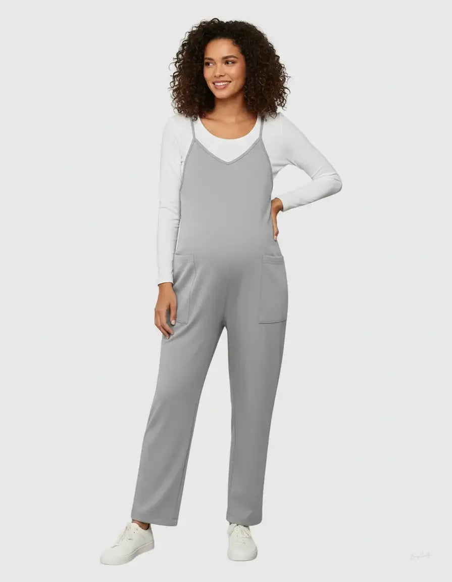 Smiling pregnant woman in a gray adjustable strap flowy maternity jumpsuit with pockets over a white long-sleeve top.