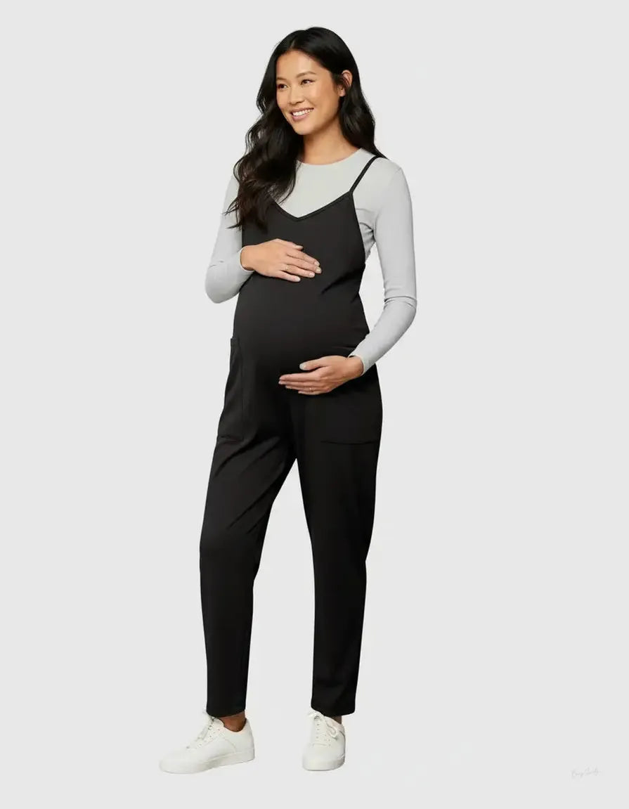 Smiling pregnant woman in a black flowy maternity jumpsuit with adjustable straps over a grey long-sleeve top.