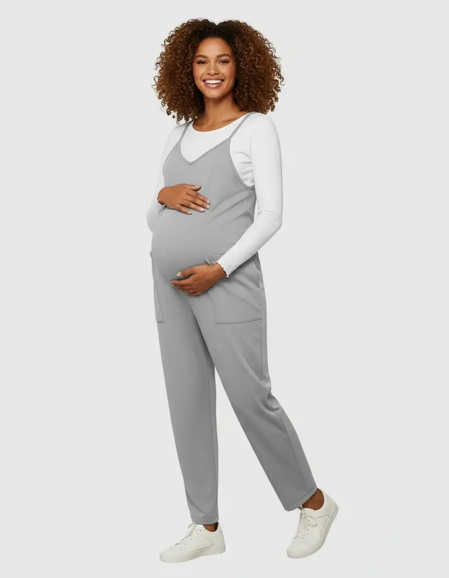 Smiling pregnant woman in a gray flowy maternity jumpsuit with adjustable straps and pockets, over a white long-sleeve top.