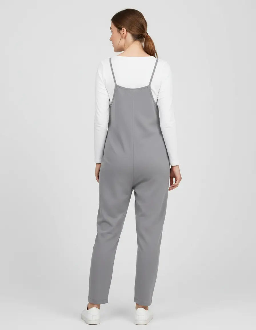 Woman in a gray flowy maternity jumpsuit with adjustable straps over a white long-sleeve shirt.
