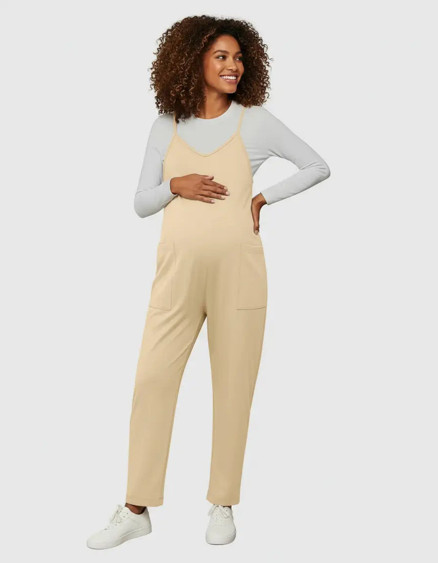 Smiling pregnant woman in a beige flowy maternity jumpsuit with adjustable straps and pockets, layered over a white long-sleeve top.