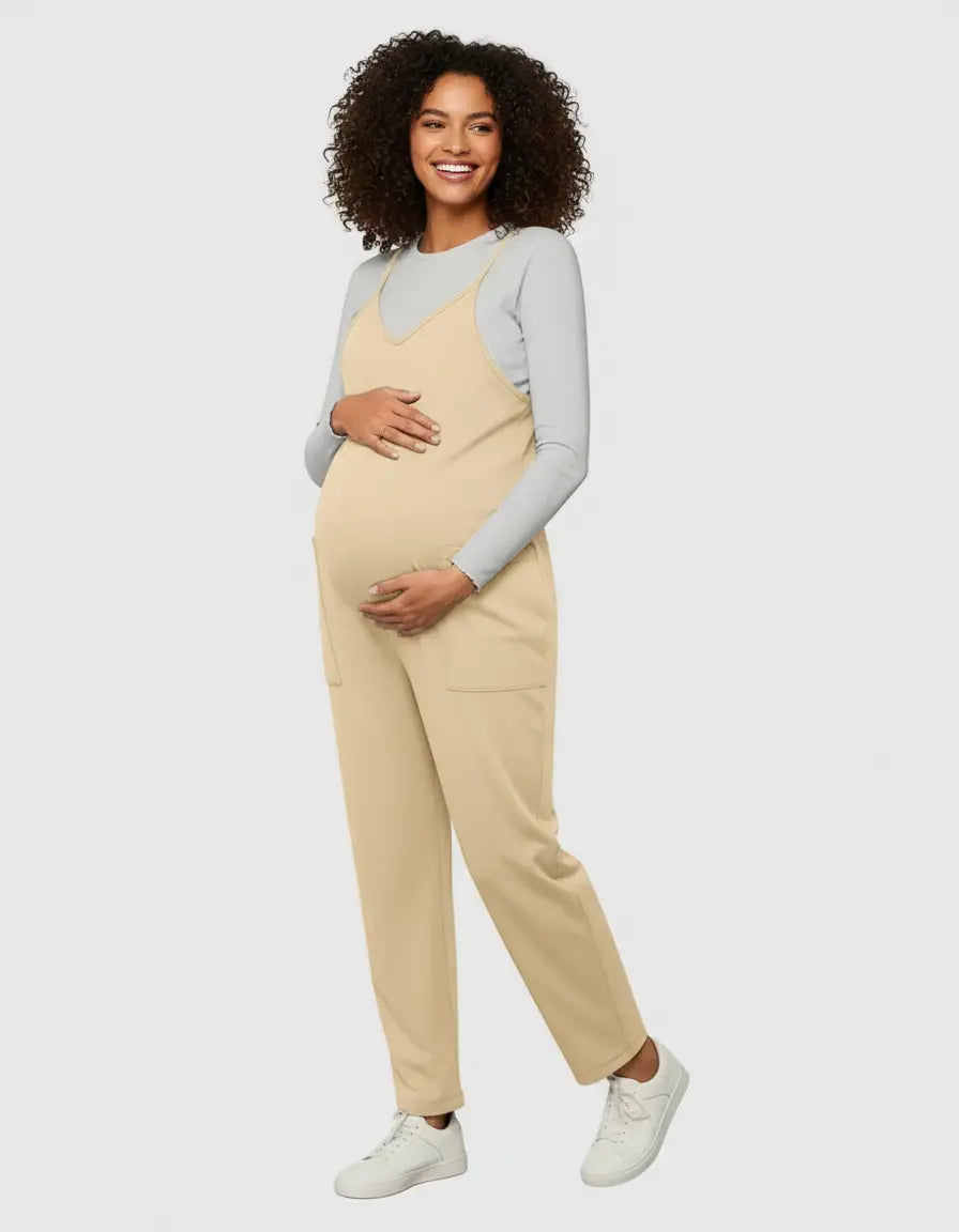 Smiling pregnant woman in a beige flowy maternity jumpsuit with adjustable straps and pockets, layered over a grey long-sleeve top.