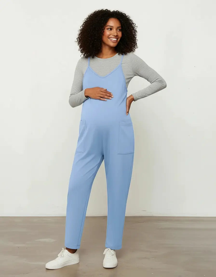 Smiling pregnant woman in a blue flowy maternity jumpsuit with adjustable straps and pockets, over a grey long-sleeve top.