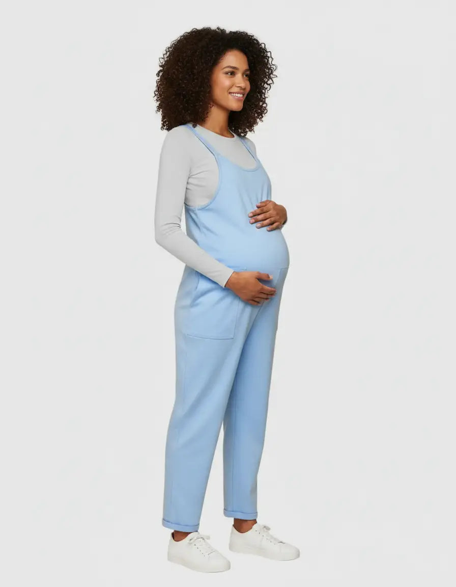 Smiling pregnant woman in a light blue flowy maternity jumpsuit with adjustable straps and pockets.