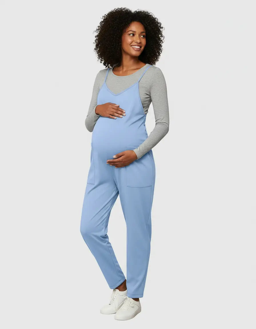 Smiling pregnant woman in a light blue flowy maternity jumpsuit with adjustable straps and pockets, over a grey long-sleeve top.