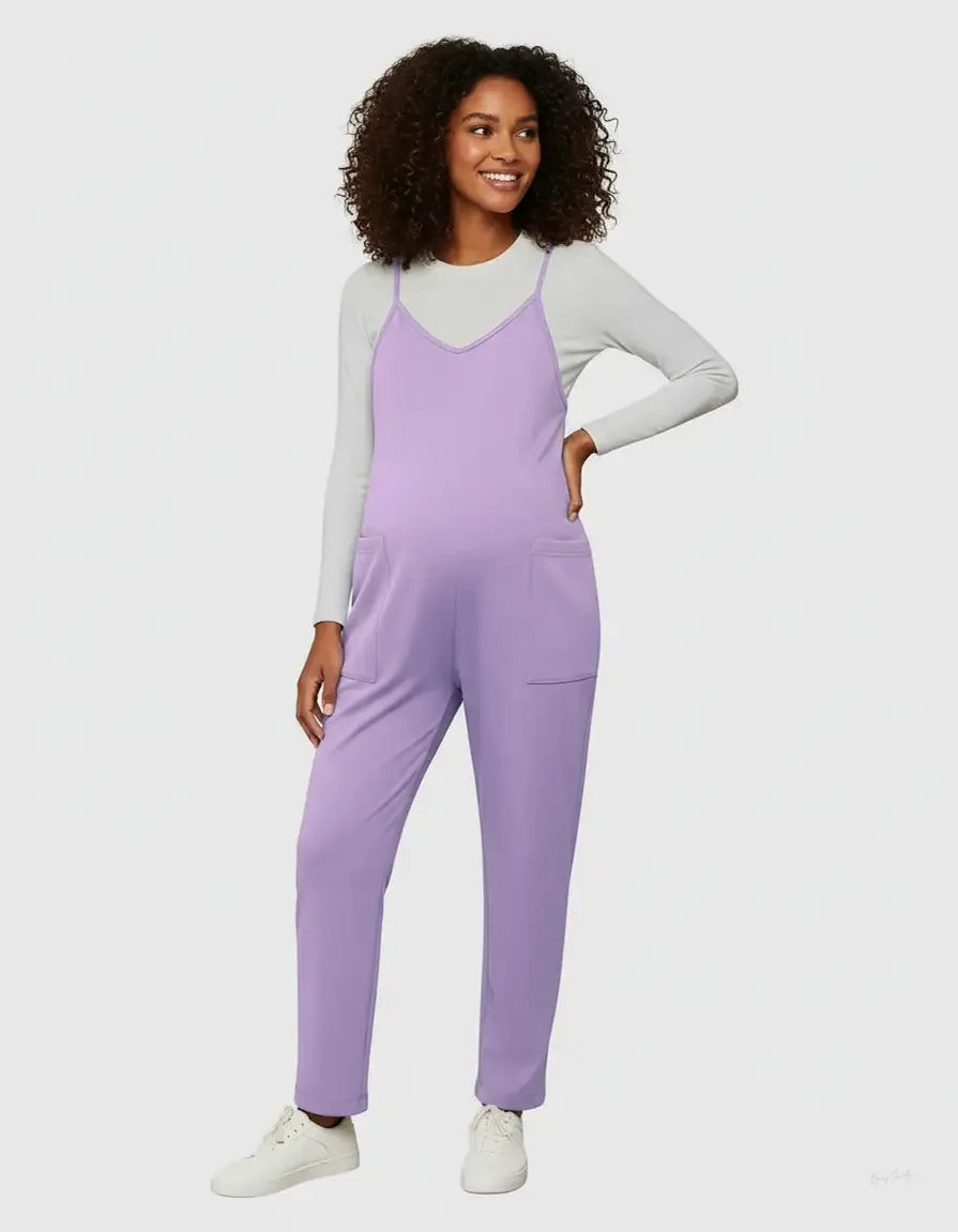 Smiling pregnant woman in a lavender flowy maternity jumpsuit with adjustable straps and pockets, over a white long-sleeve top.