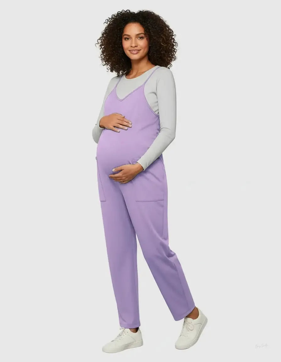 Smiling pregnant woman in a lavender flowy maternity jumpsuit with adjustable straps and pockets, over a grey long-sleeve top.