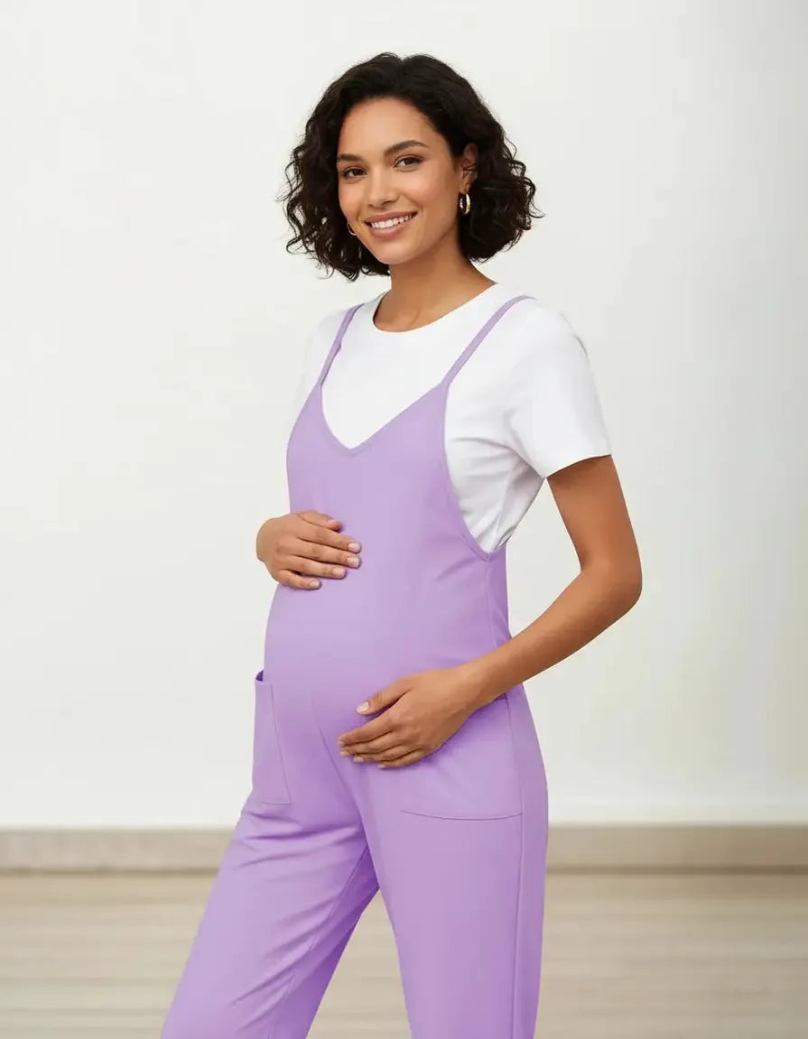 Smiling pregnant woman in a lavender flowy maternity jumpsuit with adjustable straps and pockets, over a white tee.