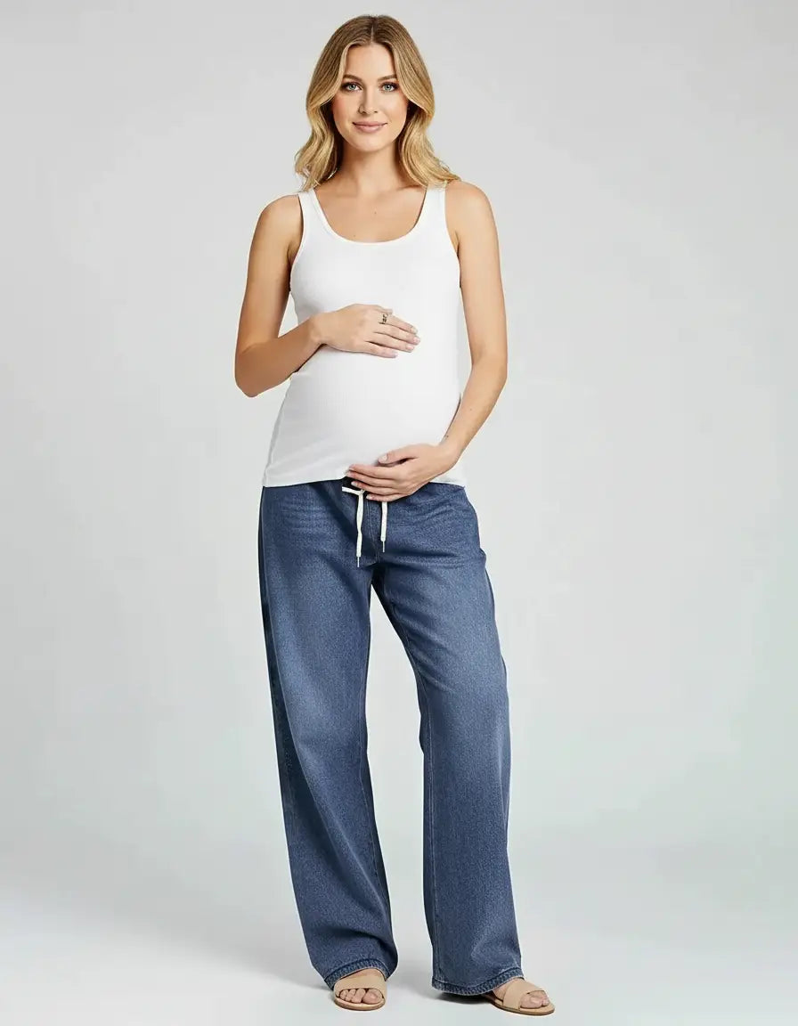 Blonde pregnant woman in a white tank top and blue wide-leg drawstring maternity jeans.