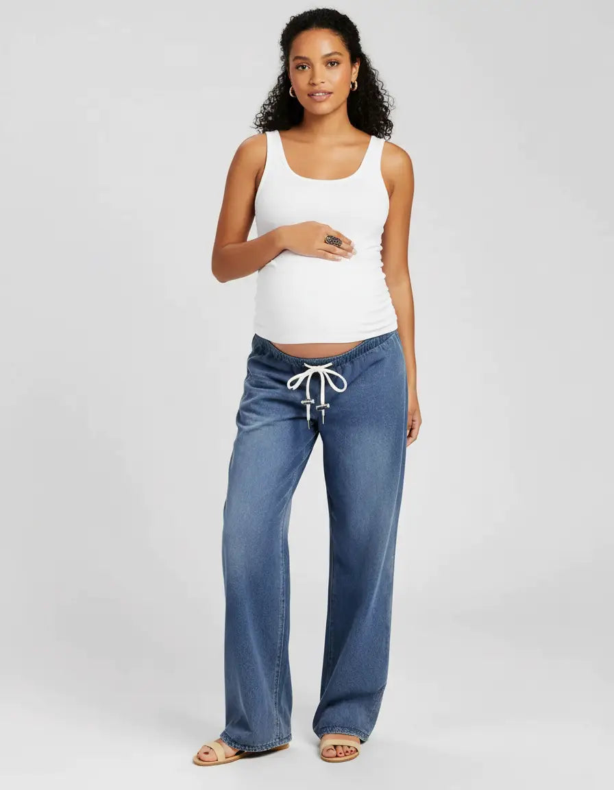 Pregnant woman in a white tank top and blue wide-leg maternity jeans with a white drawstring waist.