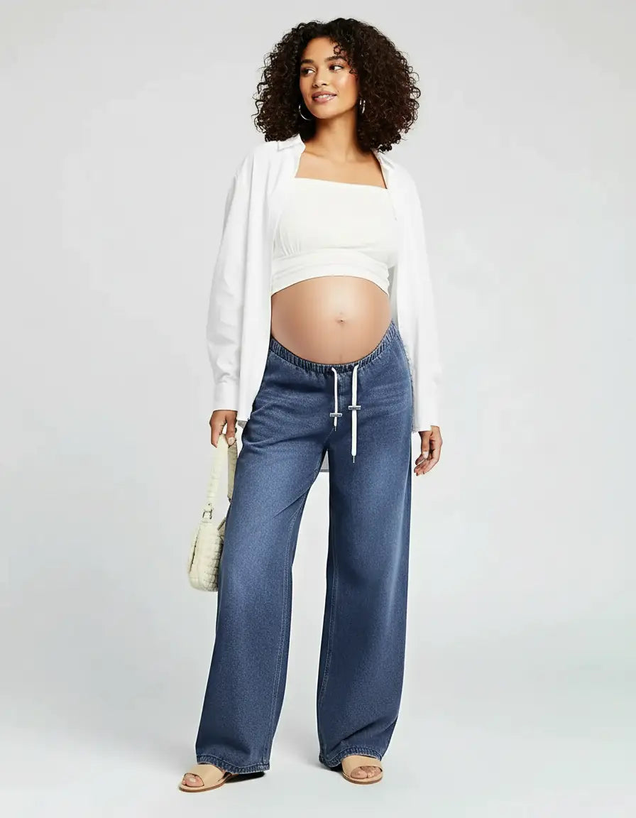 Pregnant woman in dark wash wide-leg maternity jeans with a white drawstring, white crop top, and open white shirt.