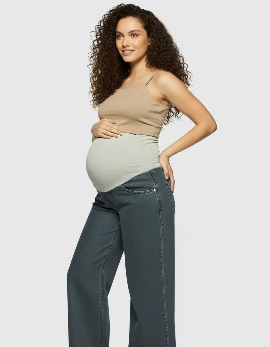 Pregnant woman in dark wash, wide-leg maternity jeans with a light green elastic waist panel and a beige tank top.