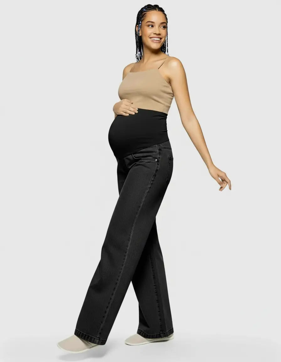 Smiling pregnant woman in black wide-leg maternity jeans with elastic waist and a beige tank top.