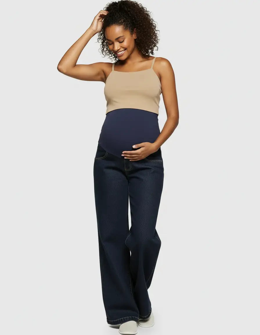 Smiling pregnant woman in a beige tank top and dark wash wide-leg maternity jeans with a navy elastic waist.